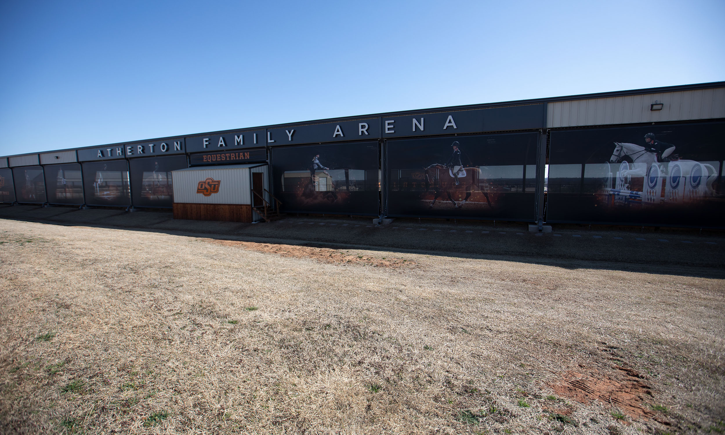 The building has a long facade with large windows displaying images of equestrian activities. There is a sign with the OSU logo near the entrance. The ground in front of the arena appears to be dry and grassy.