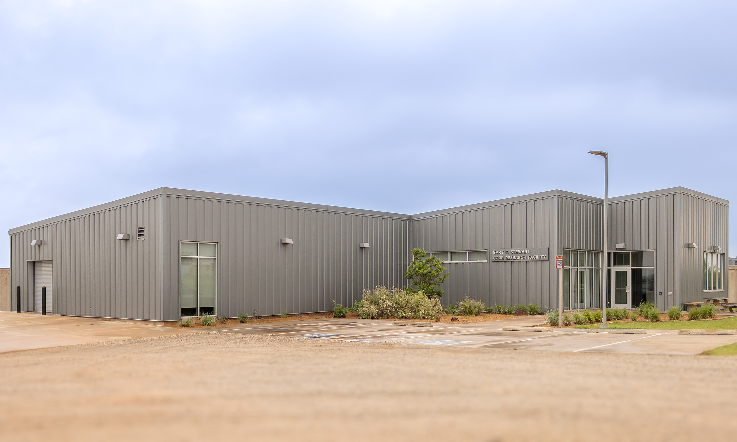 The image depicts a large industrial building with corrugated metal siding and flat roofs with a large parking lot.