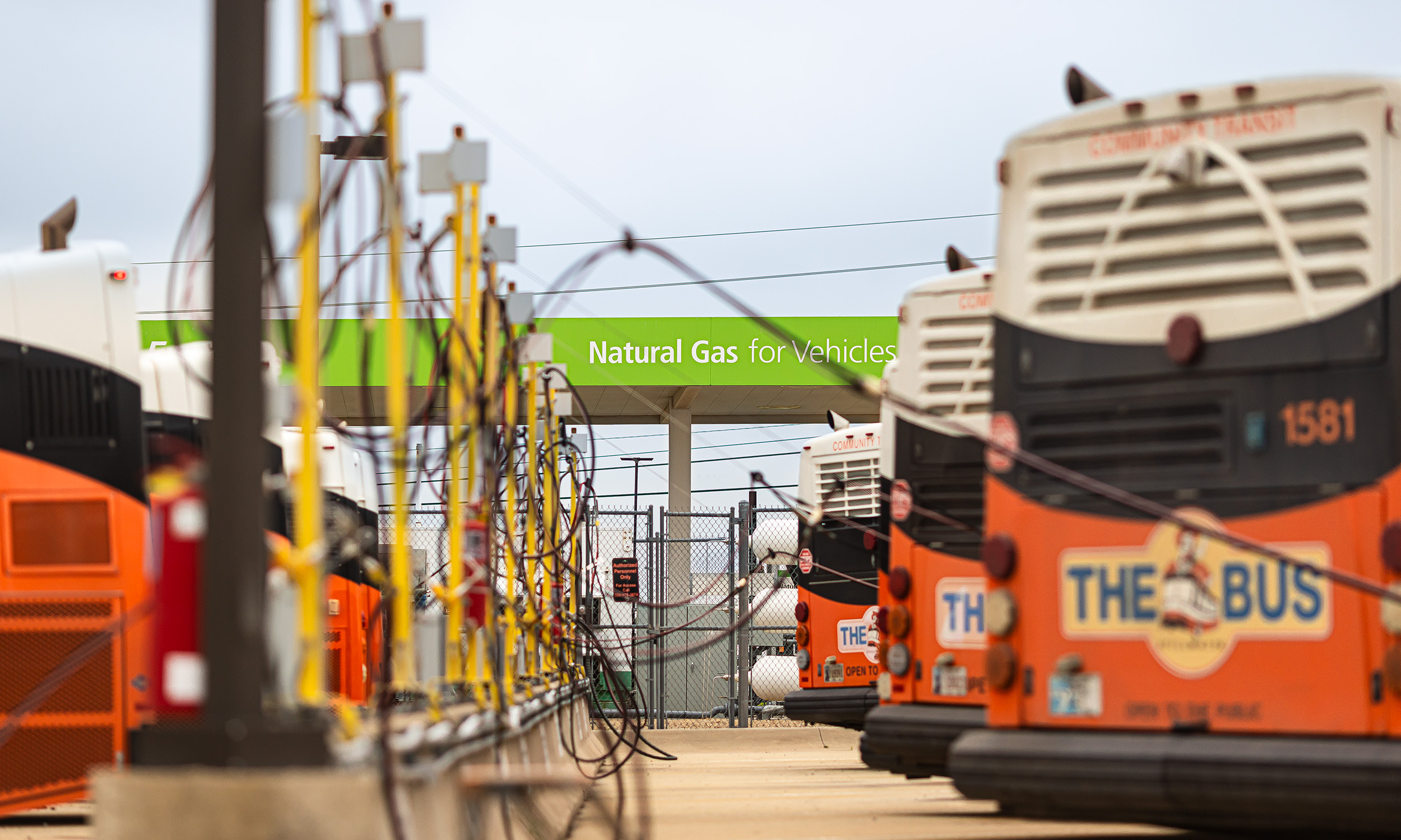The image shows a bus depot with several buses parked in rows. The buses are connected to fueling stations with hoses, indicating that they are being refueled. A green sign in the background reads "Natural Gas for Vehicles," suggesting that the buses run on natural gas.