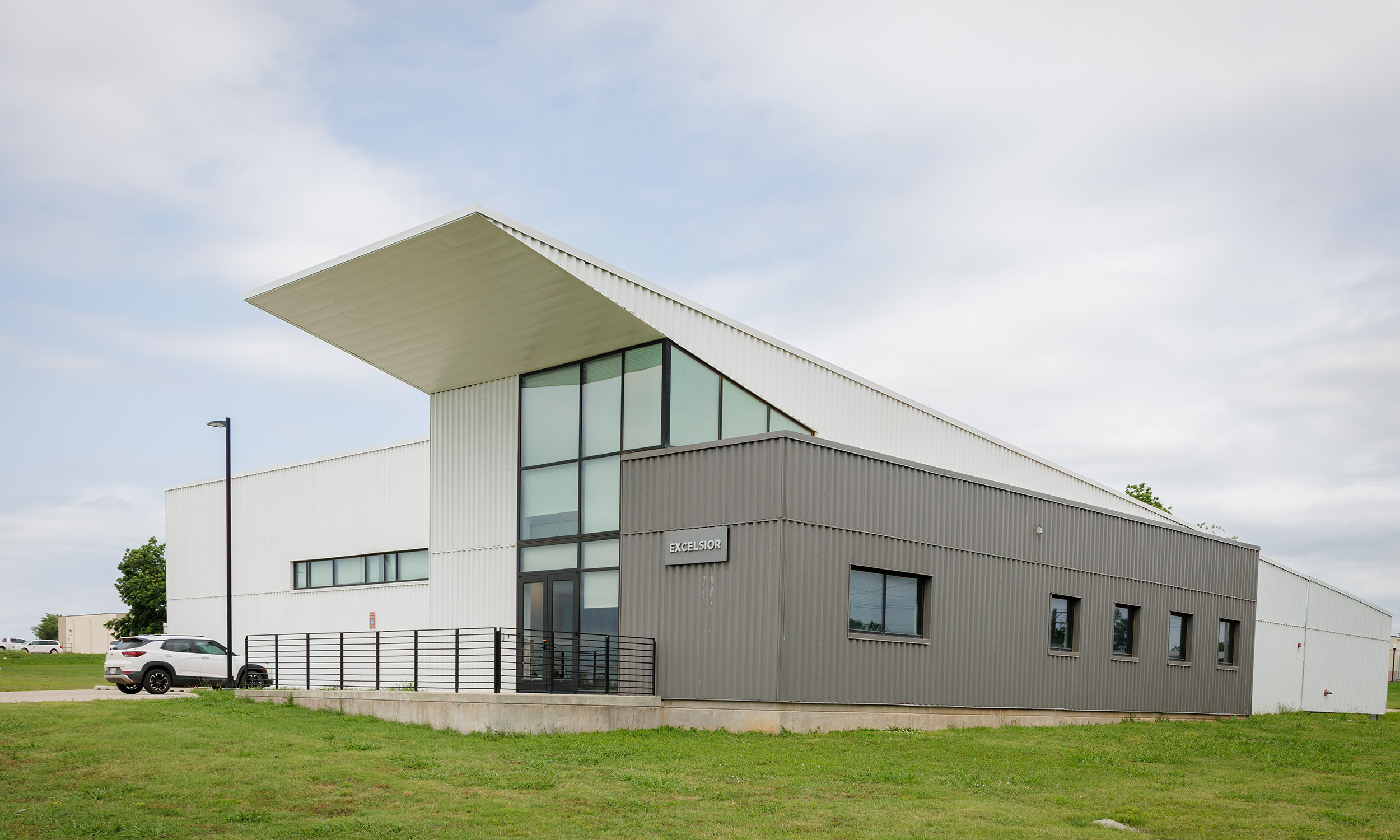 A modern building with a unique angular roof design, featuring large glass windows and a combination of white and gray exterior panels. The building has a small entrance area with a ramp and railing. 
