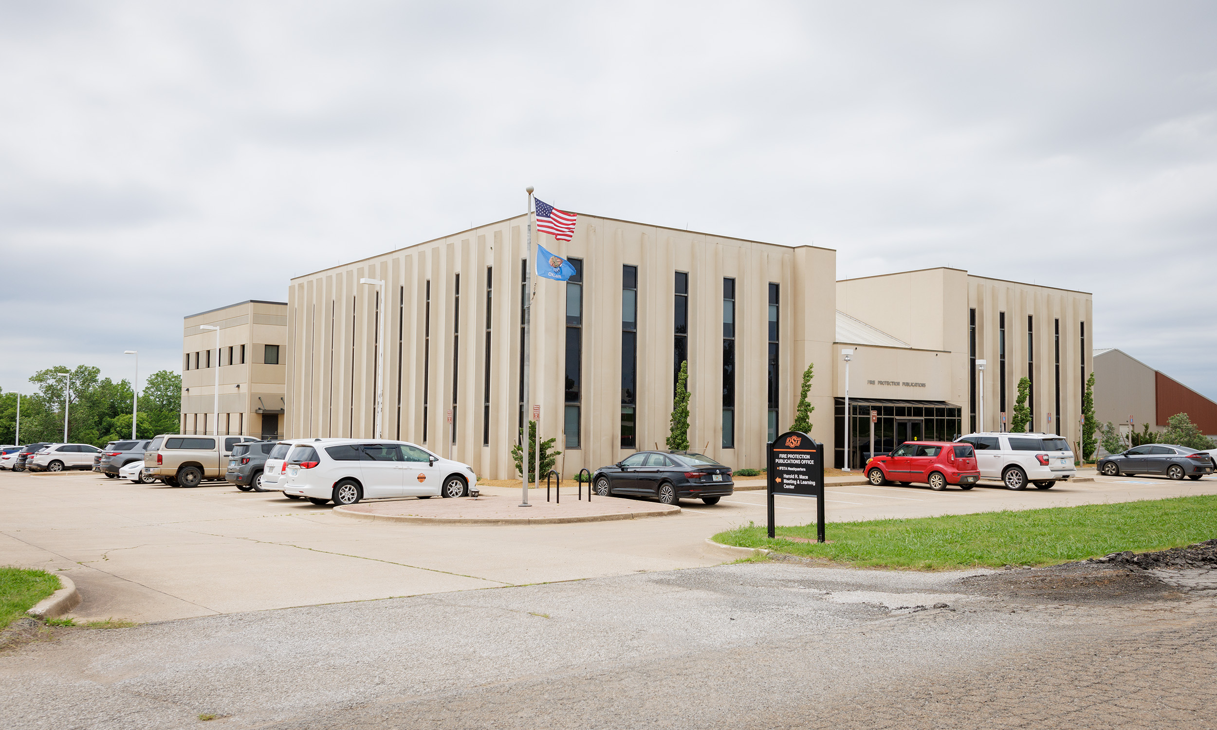 A modern, beige-colored office building with vertical windows. Several cars are parked in the parking lot in front of the building. An American flag and another flag are flying on a flagpole near the entrance