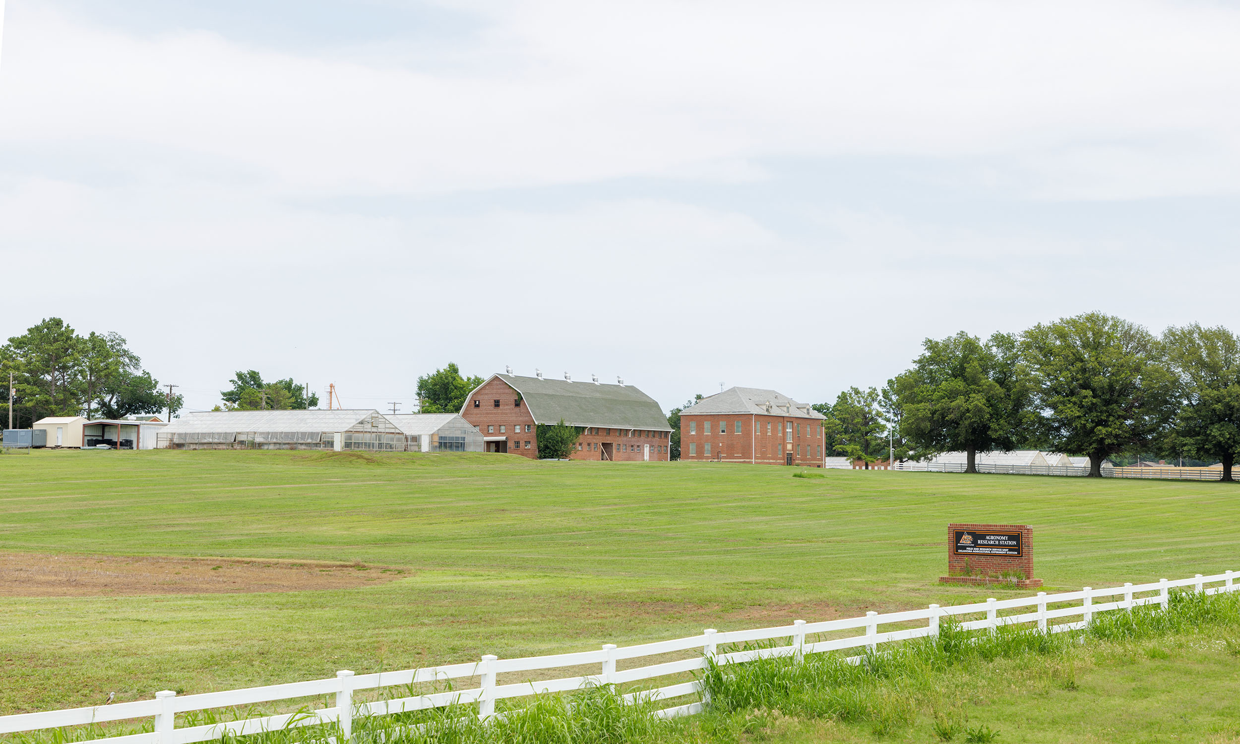 The image shows a wide, open field with a white fence in the foreground. In the middle distance, there are several buildings, including greenhouses and barns.