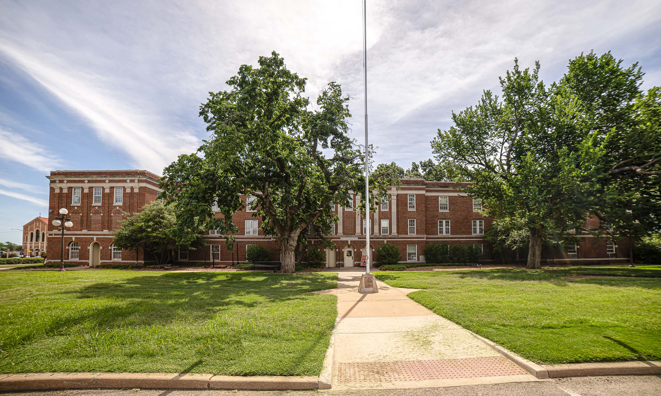 A large, three-story brick building with white trim and numerous windows, surrounded by a well-maintained lawn and mature trees. A flagpole stands in the center of a paved walkway leading to the building's entrance, under a partly cloudy sky.