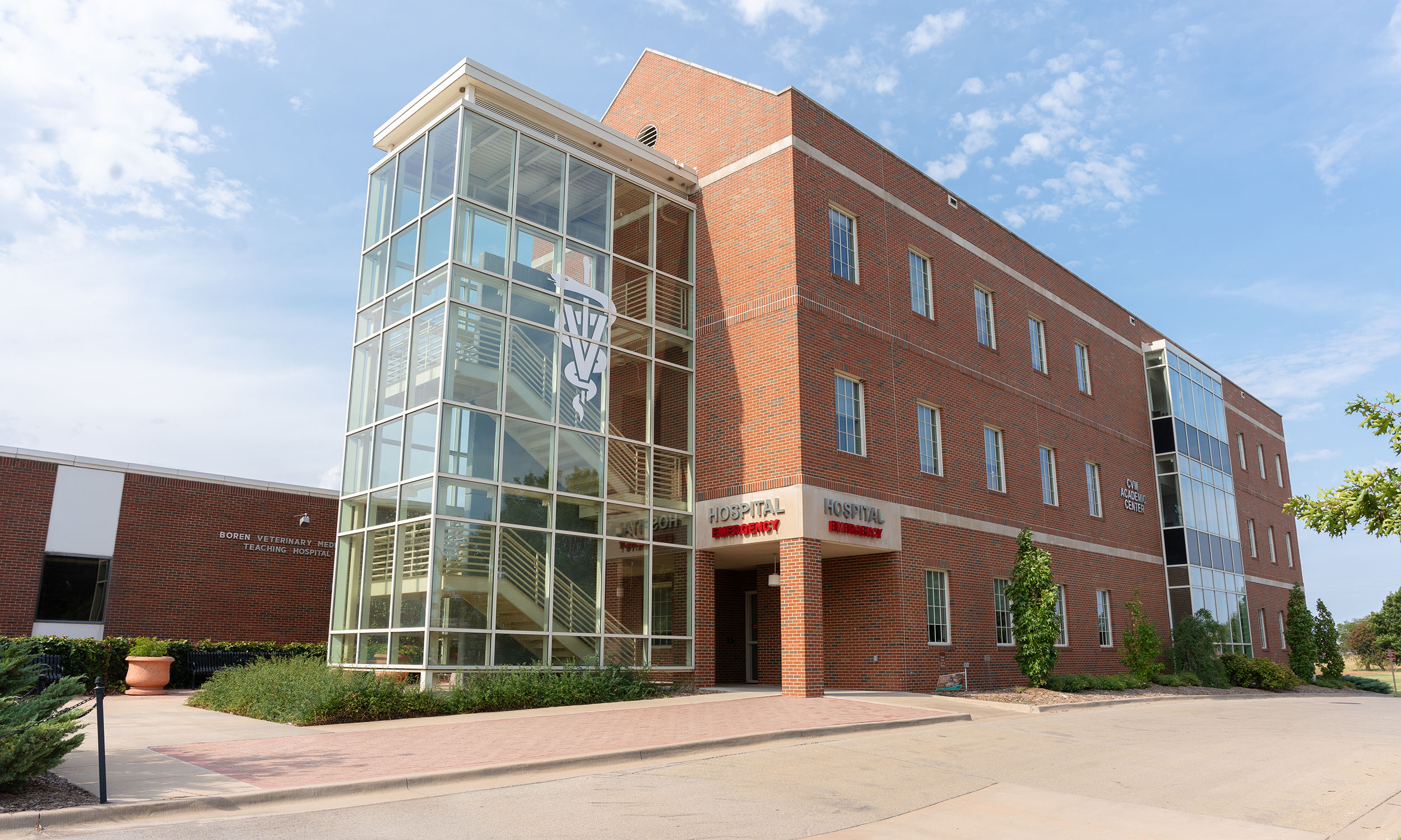 A three-story brick building with a glass-enclosed stairwell on the left side. A sign reading 'HOSPITAL' is displayed above the entrance, which is located under a small overhang. The building is surrounded by plants and shrubs, with a clear sky and a few clouds overhead.