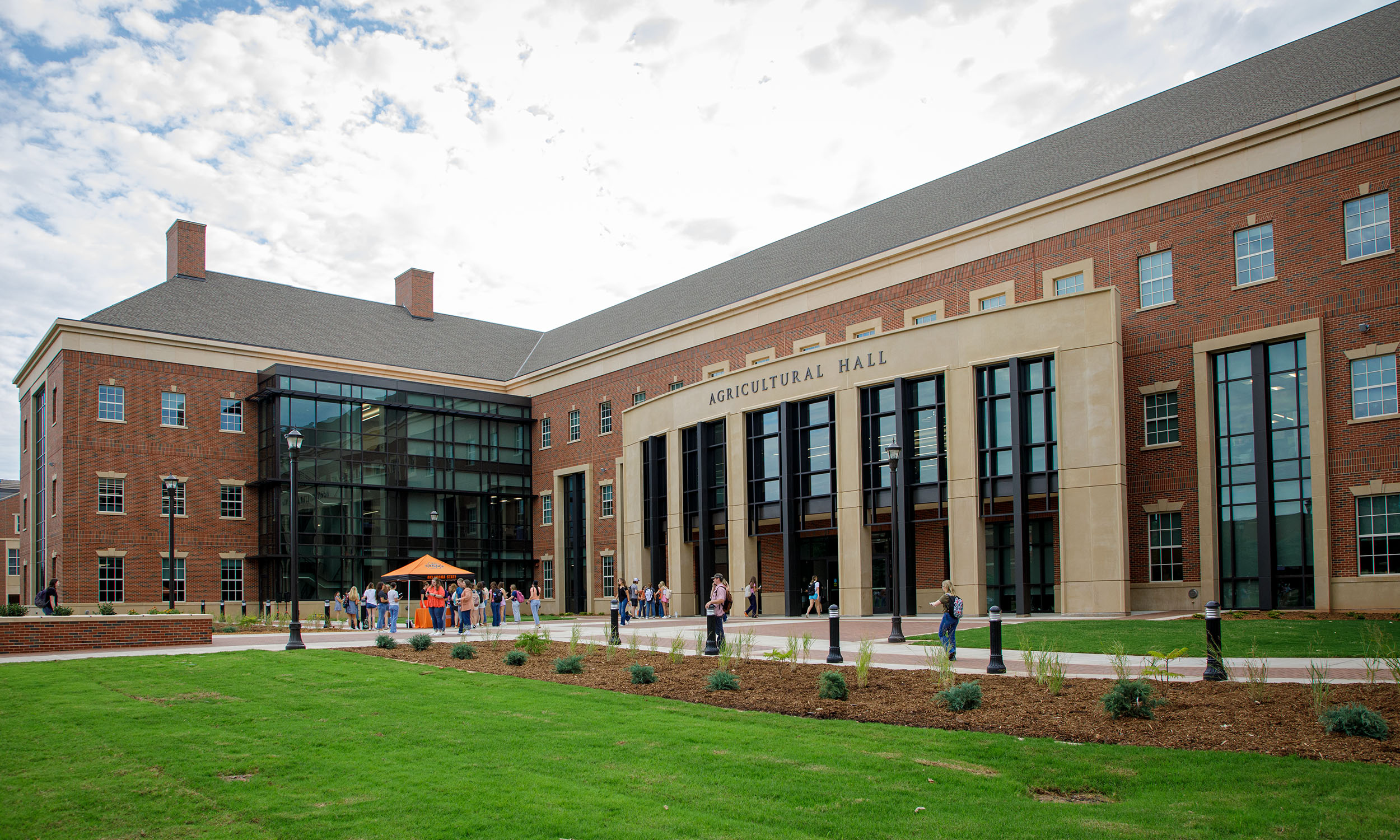 A large brick building with a modern glass extension, labeled 'Agricultural Hall,' shown on a sunny day. The building has multiple stories and features tall windows. In front of the building, there is a well-maintained lawn with some landscaping and several people walking or standing near the entrance.