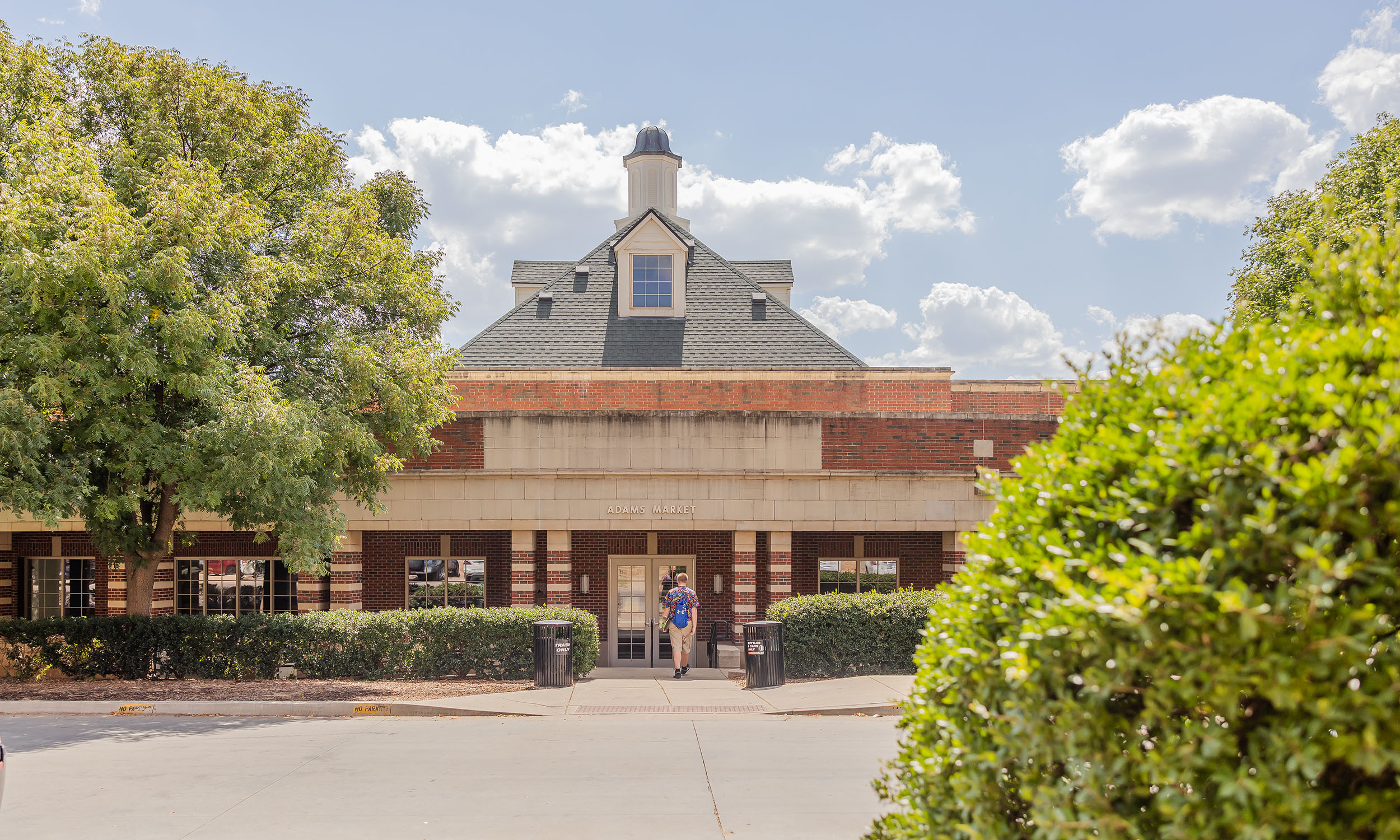 Exterior view of Adams Market building on a sunny day, with a person walking out the front entrance. The building features red brick, white stone accents, and a pitched roof with dormer windows. Trees and bushes frame the scene under a partly cloudy sky.