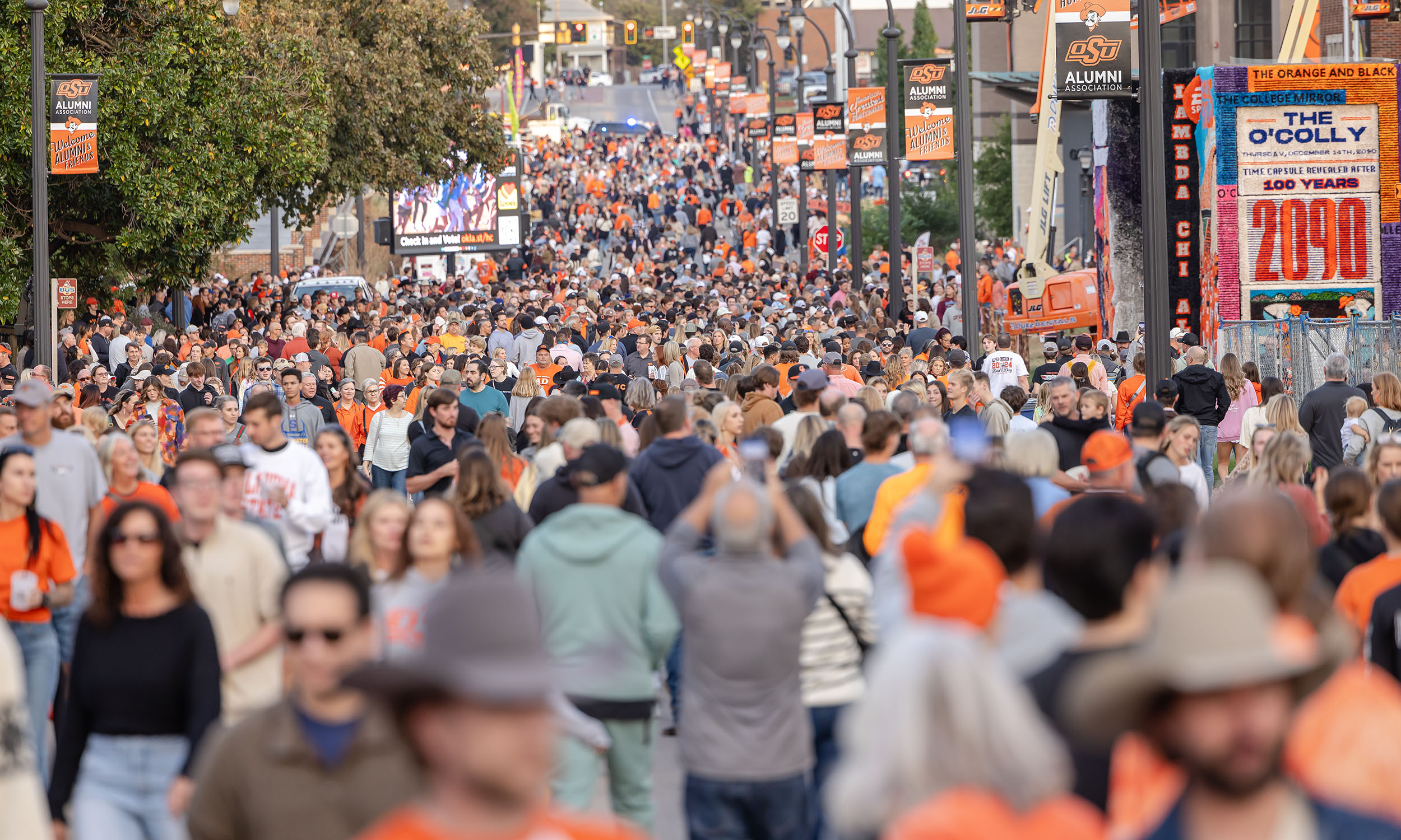 A large crowd of people, many wearing orange clothing, walk down a street lined with trees and banners. The street is filled with people, and there are signs and advertisements visible in the background.