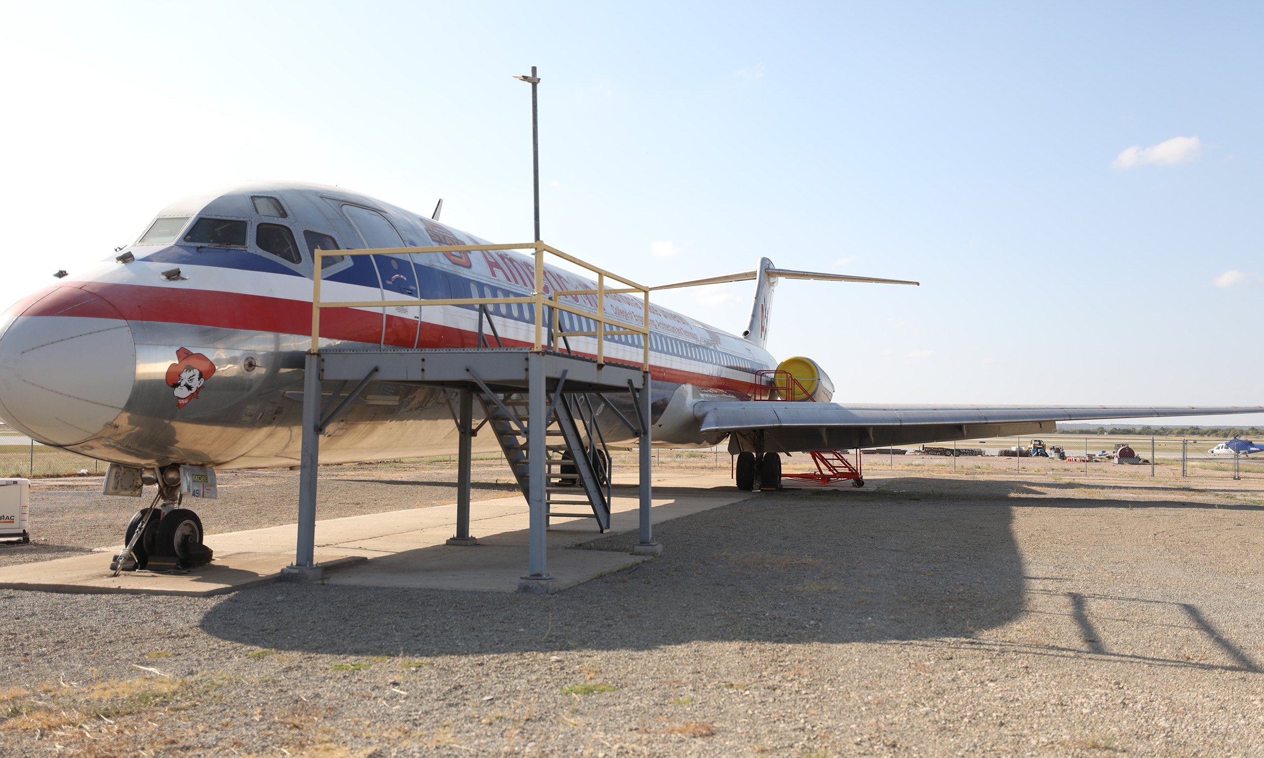 An old American Airlines aircraft is parked on a gravel surface. The plane has a red, white, and blue stripe running along its fuselage. A set of stairs with yellow railings leads up to the aircraft's door. The tail section of the plane is visible in the background, and there are some scattered objects on the ground nearby. The sky is clear with a few clouds.