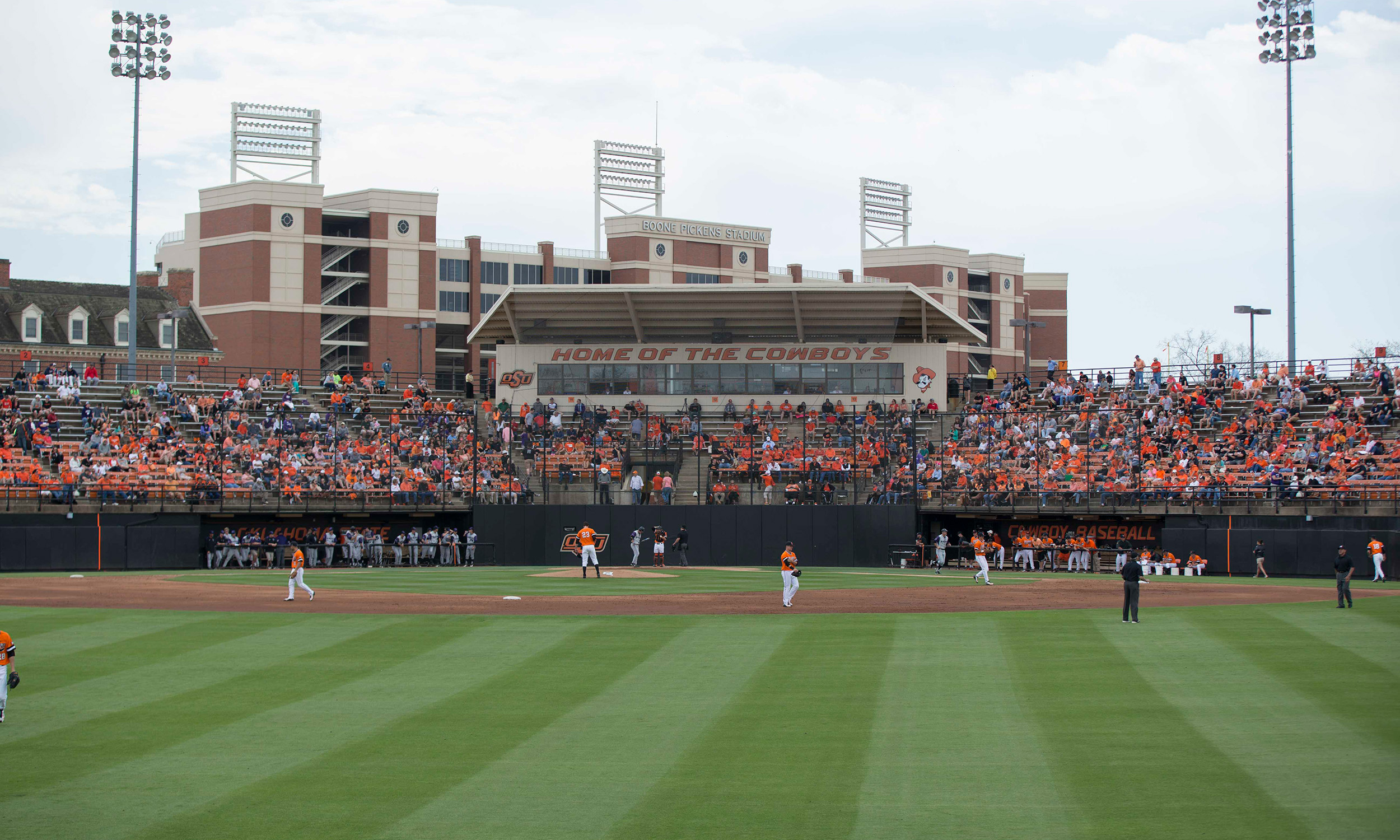 A baseball stadium filled with spectators, with a large building in the background displaying the sign 'Home of the Cowboys.' The field is well-maintained with green grass, and players are visible on the field. The stands are packed with fans wearing orange and black, indicating support for the home team. The sky is partly cloudy, and there are tall light poles around the stadium.