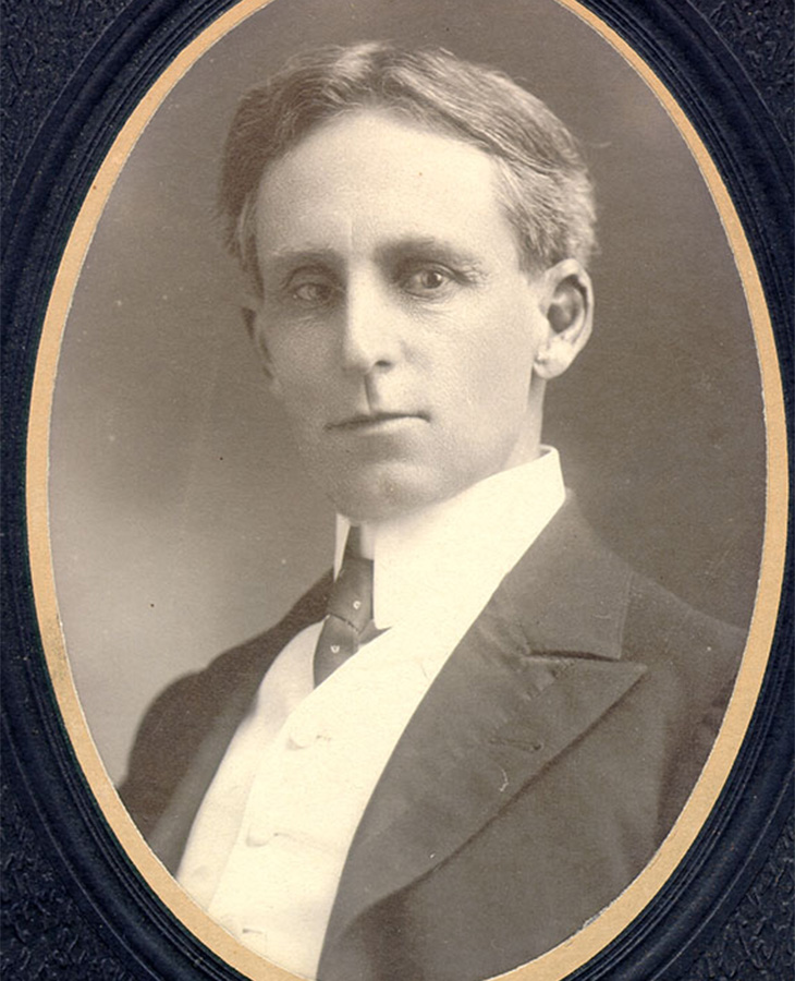 A sepia-toned portrait of a man wearing a formal suit with a high-collared shirt and tie. The man has short, neatly combed hair and is framed within an oval border.