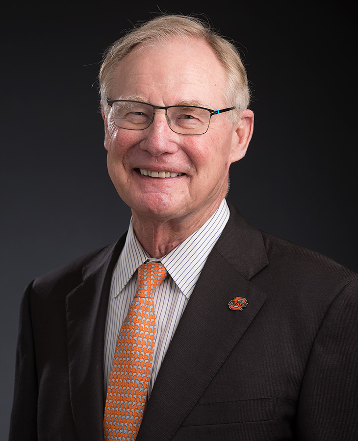 A person wearing a dark suit jacket with an Oklahoma State University lapel pin, a white dress shirt with thin vertical stripes and an orange patterned tie.