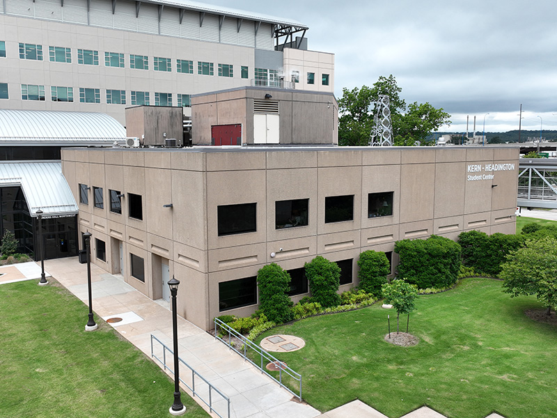 A two-story beige building with several windows. In front is a well-maintained lawn with bushes and trees, a sidewalk and street lamps. A larger multi-story building and industrial structures are visible in the background.