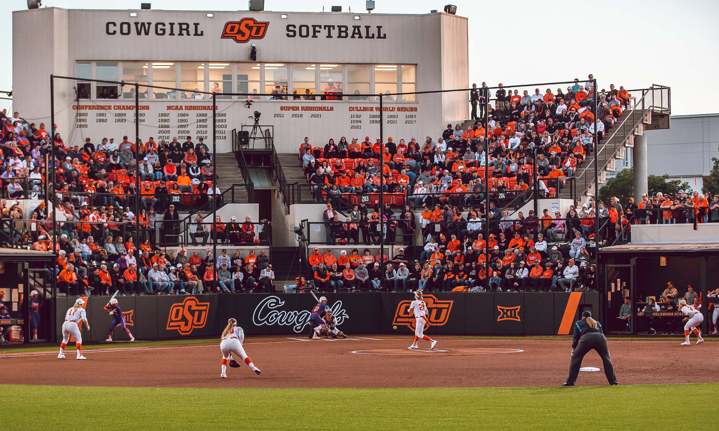 A softball game is taking place at Cowgirl Stadium, home of the Oklahoma State University (OSU) Cowgirl Softball team. The stadium is filled with spectators, many wearing orange shirts. On the field, one player is pitching the ball while others are positioned defensively.
