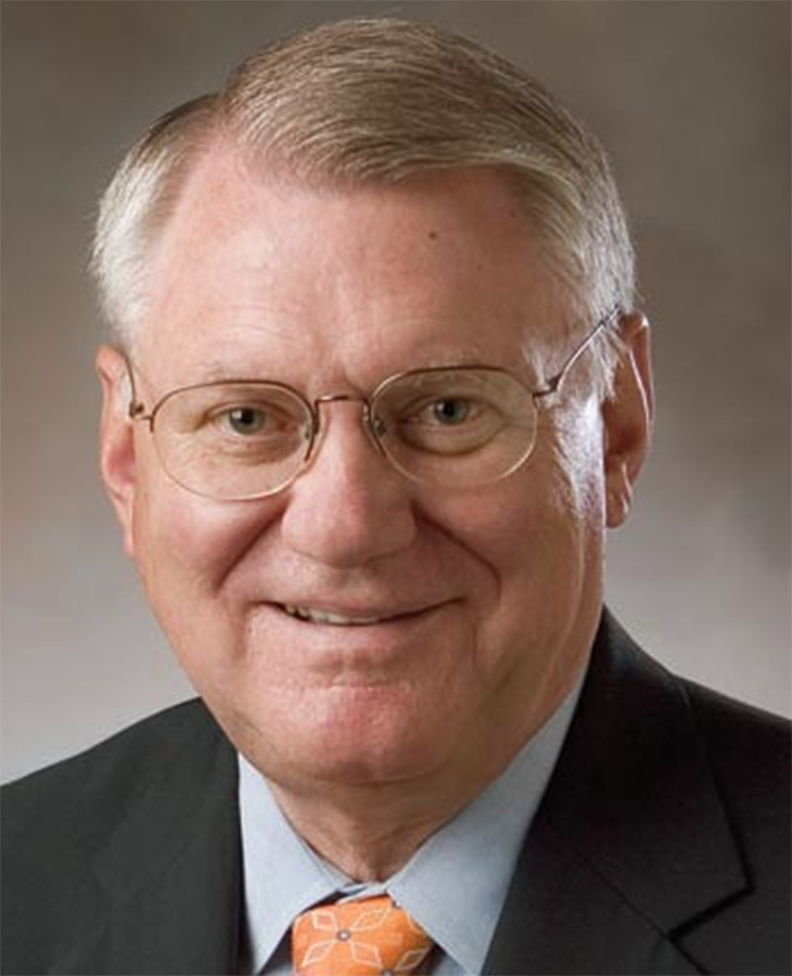 An older man with gray hair wearing glasses, a dark suit, a white shirt, and an orange patterned tie. The background is plain and neutral.