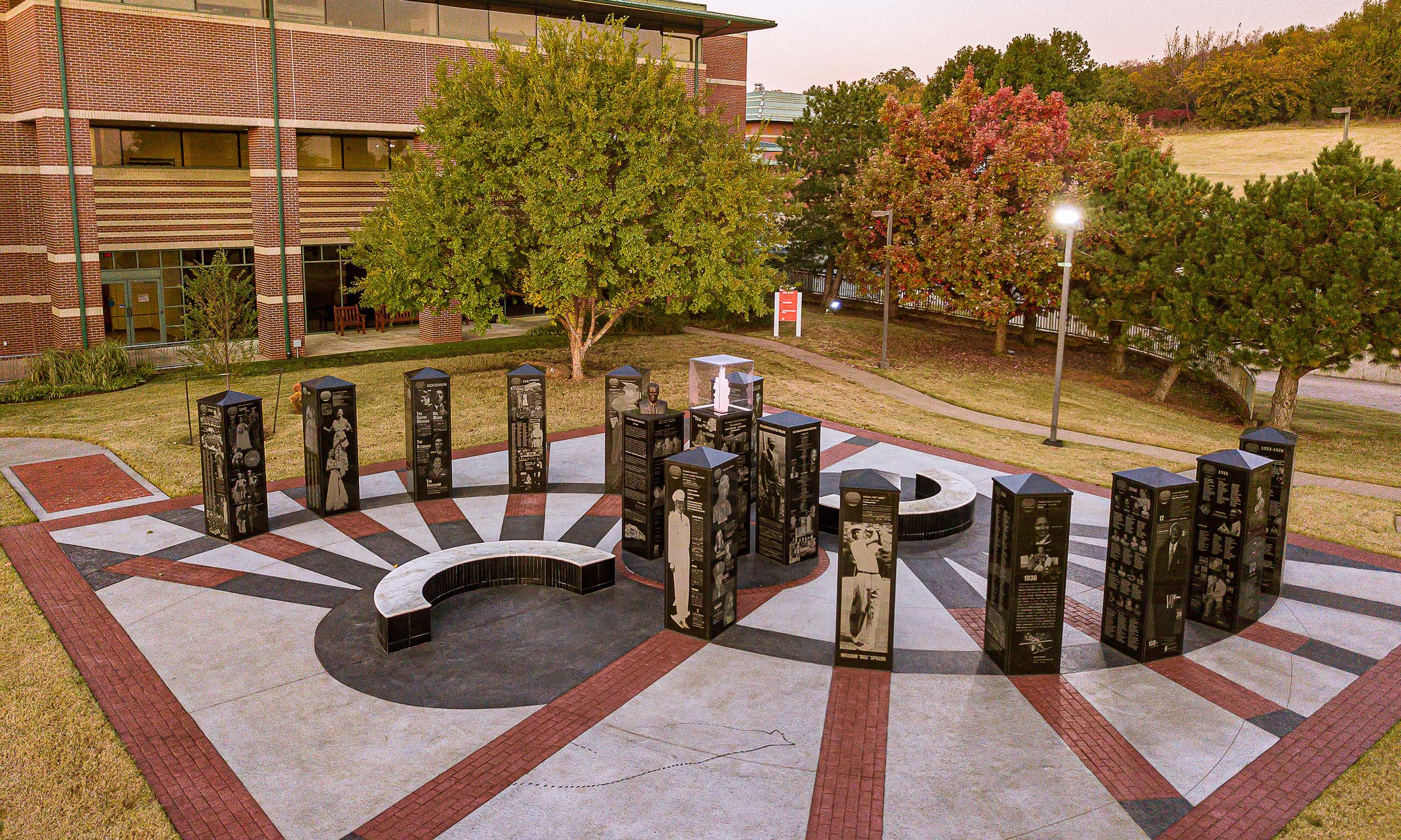 An outdoor memorial with multiple black granite pillars arranged in a semi-circle on a patterned concrete surface. Each pillar has engraved images and text. The memorial is surrounded by trees with autumn foliage, and a building is visible in the background.