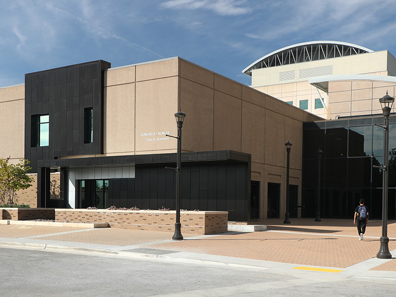 A modern beige building with black accents, large windows and a flat roof. Street lamps surround the building and a person is walking on the sidewalk in front. The sky is clear with some clouds.