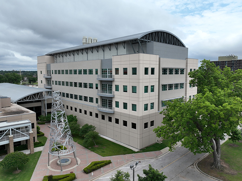 A large multi-story beige building with many windows and a curved roof. In front is a modern metal sculpture surrounded by a circular pathway and landscaped greenery. A tree is on the right and part of another building is visible on the left under a cloudy sky with blue patches