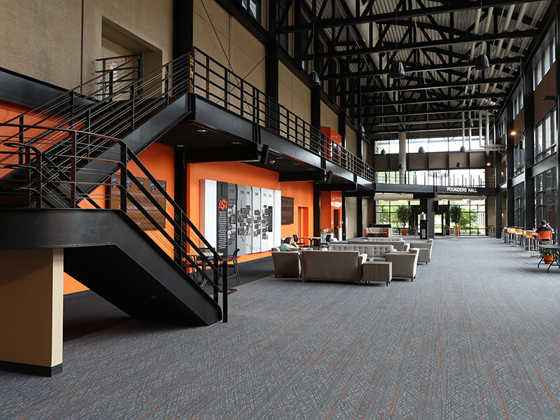 A spacious modern lobby with high ceilings and large windows. A black metal staircase with orange walls underneath is on the left. Gray couches and chairs form a seating area in the center. 