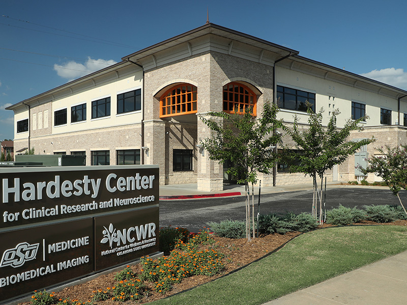A two-story beige brick building with large windows. In front is a sign reading 'Hardesty Center for Clinical Research and Neuroscience' with logos for OSU Medicine, Biomedical Imaging and NCWR. The building is surrounded by grass, shrubs and flowers.