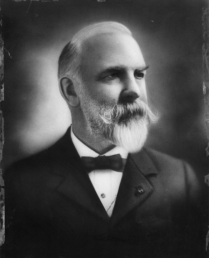 A black and white portrait of an elderly man. The man is wearing a formal suit with a bow tie. The edges of the photograph are worn and slightly damaged, giving it an antique appearance.