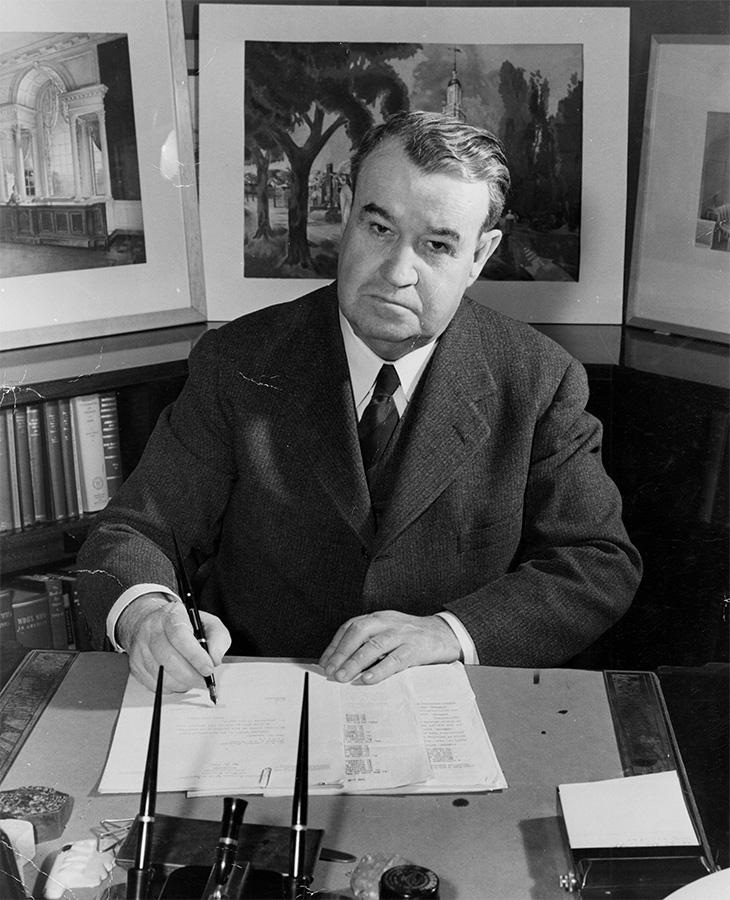 A man is sitting at a desk, wearing a suit and tie. He is writing on a document with a pen. Behind him are framed pictures of architectural scenes and trees. The desk has various items including an ink bottle, pens, and papers.