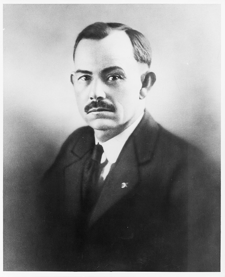 A black and white portrait photograph of a man wearing a dark suit jacket, white shirt, and a dark tie. The background is plain and light-colored.