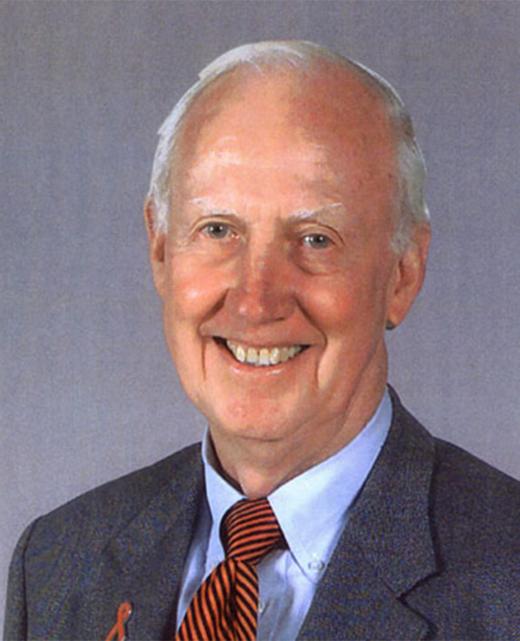 An older person with white hair wearing a gray suit, white shirt, and a red and black striped tie. An orange ribbon is pinned to the left lapel of the suit jacket.