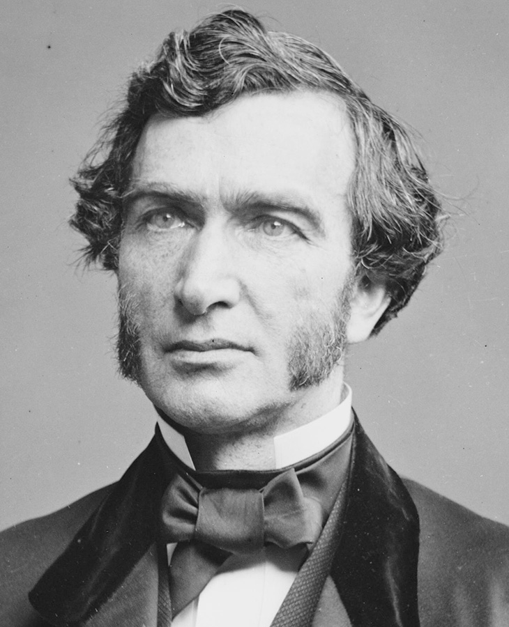 A person with medium-length, wavy hair and a beard, dressed in a formal suit with a bow tie, posed against a plain gray background.
