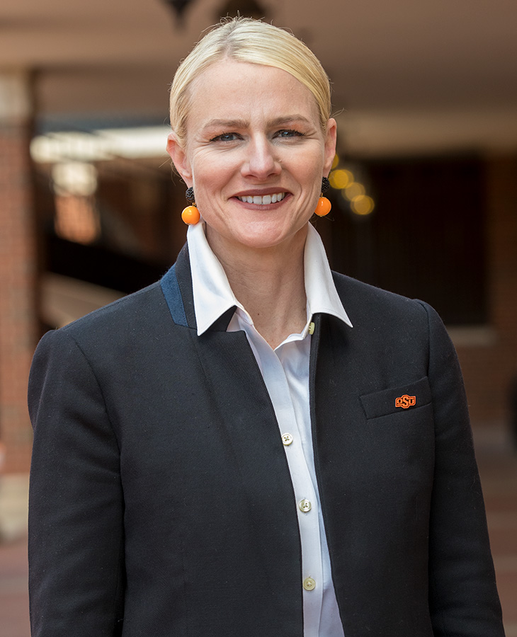 A woman with blonde hair wearing a white collared shirt under a dark blazer that features an orange and black logo on the left chest.