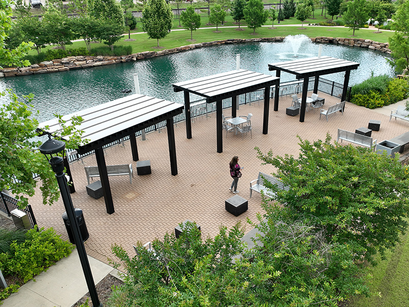 An outdoor park with a paved walkway and several covered seating areas featuring black metal frames and white roofs. Benches and tables are under the shelters. 