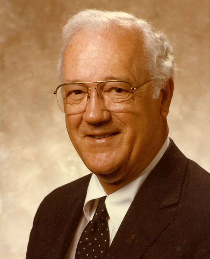 An older man with white hair is wearing a dark suit, white shirt, and a black tie with small white polka dots. The background is a neutral beige color. 