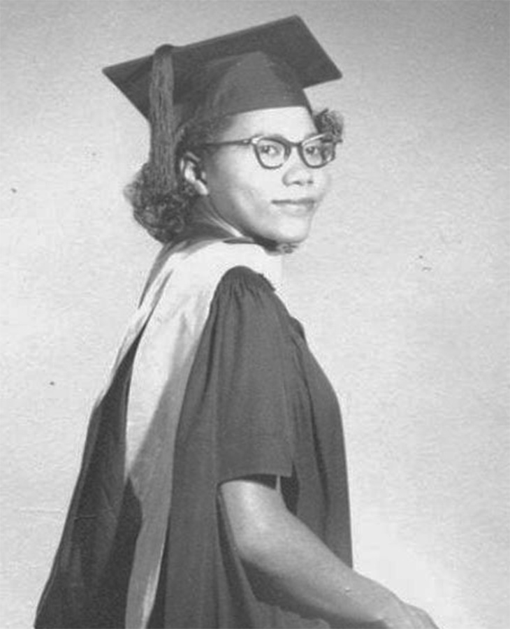 A woman wearing a graduation cap and gown, viewed from the side. She has shoulder-length curly hair and is standing against a plain background.