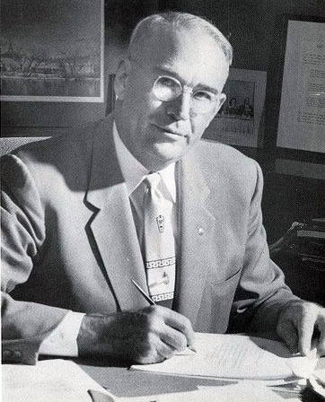 An older man in a suit and tie is sitting at a desk, holding a pen and writing on papers. The background includes framed pictures and documents on the wall.