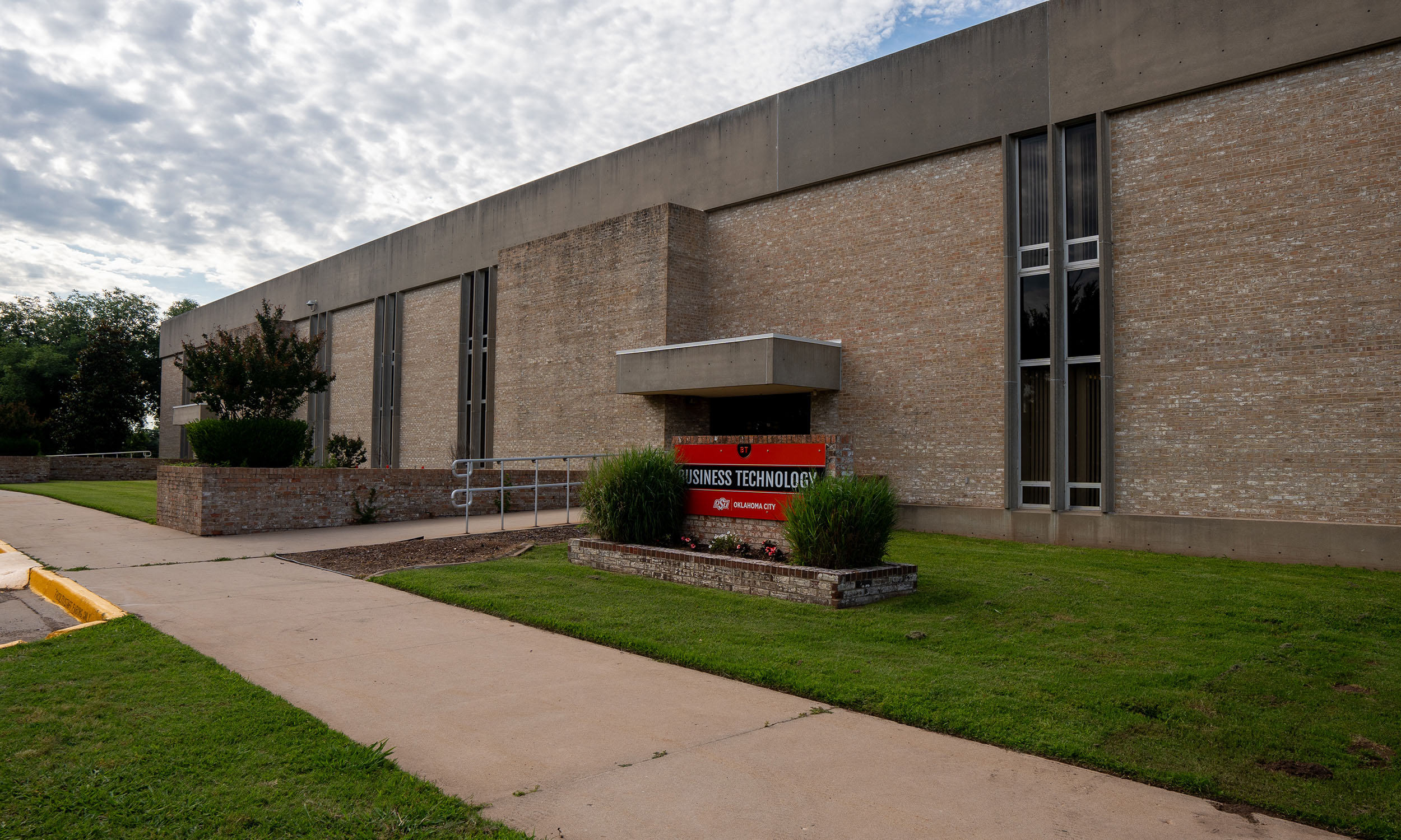 A single-story brick building with a concrete roof and large vertical windows. In front of the building, there is an orange sign that reads 'Business Technology'