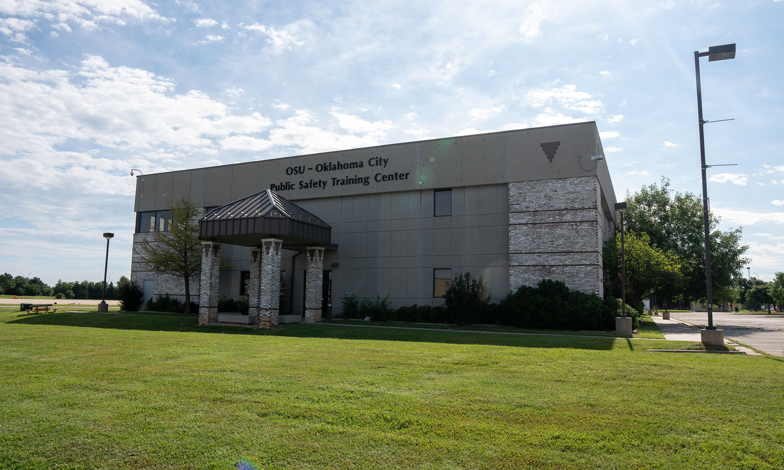 A modern building labeled 'OSU - Oklahoma City Public Safety Training Center' with a brick and concrete exterior. A small covered entrance supported by columns is at the front, surrounded by a neatly maintained lawn and trees under a clear sky.