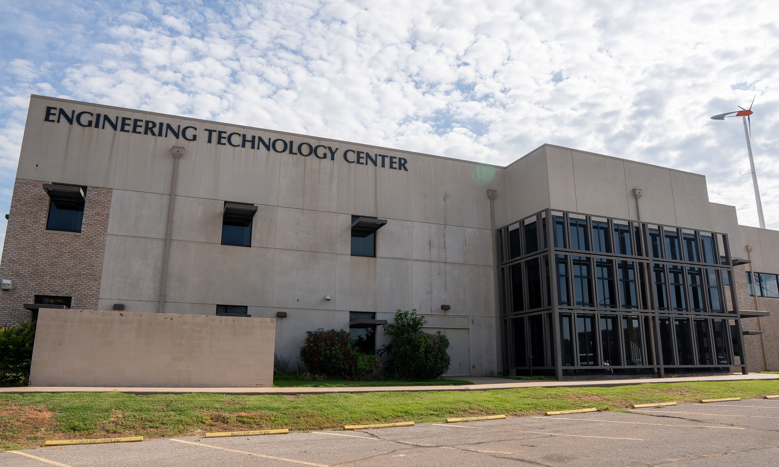 A two-story building with a sign that reads 'ENGINEERING TECHNOLOGY CENTER' on the upper part of the facade. The building has a mix of brick and concrete exterior, with large windows on the right side. 