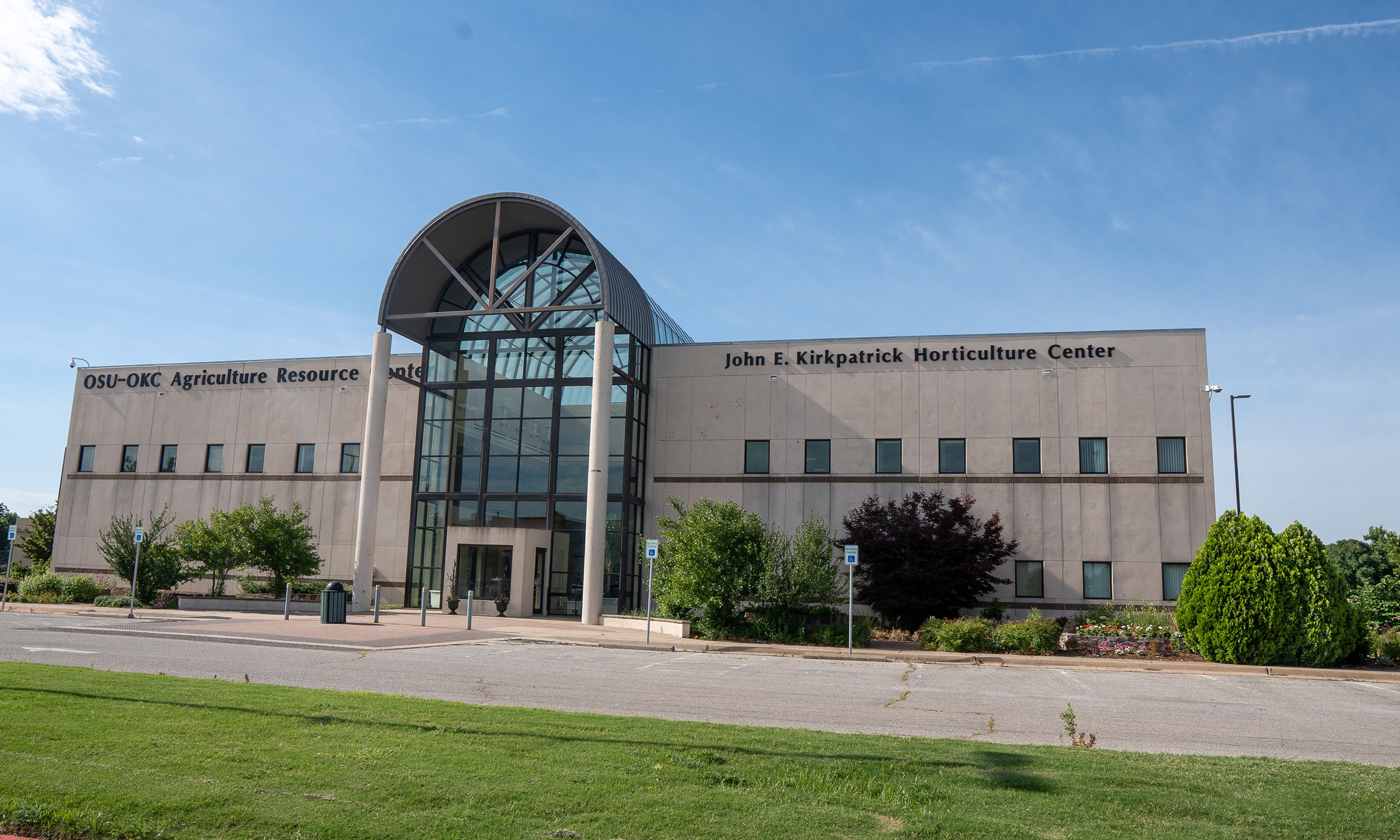 A large beige building with a glass atrium entrance and the words 'OSU-OKC Agriculture Resource Center' and 'John E. Kirkpatrick Horticulture Center' written on the facade. The building is surrounded by a well-maintained lawn, bushes and trees.