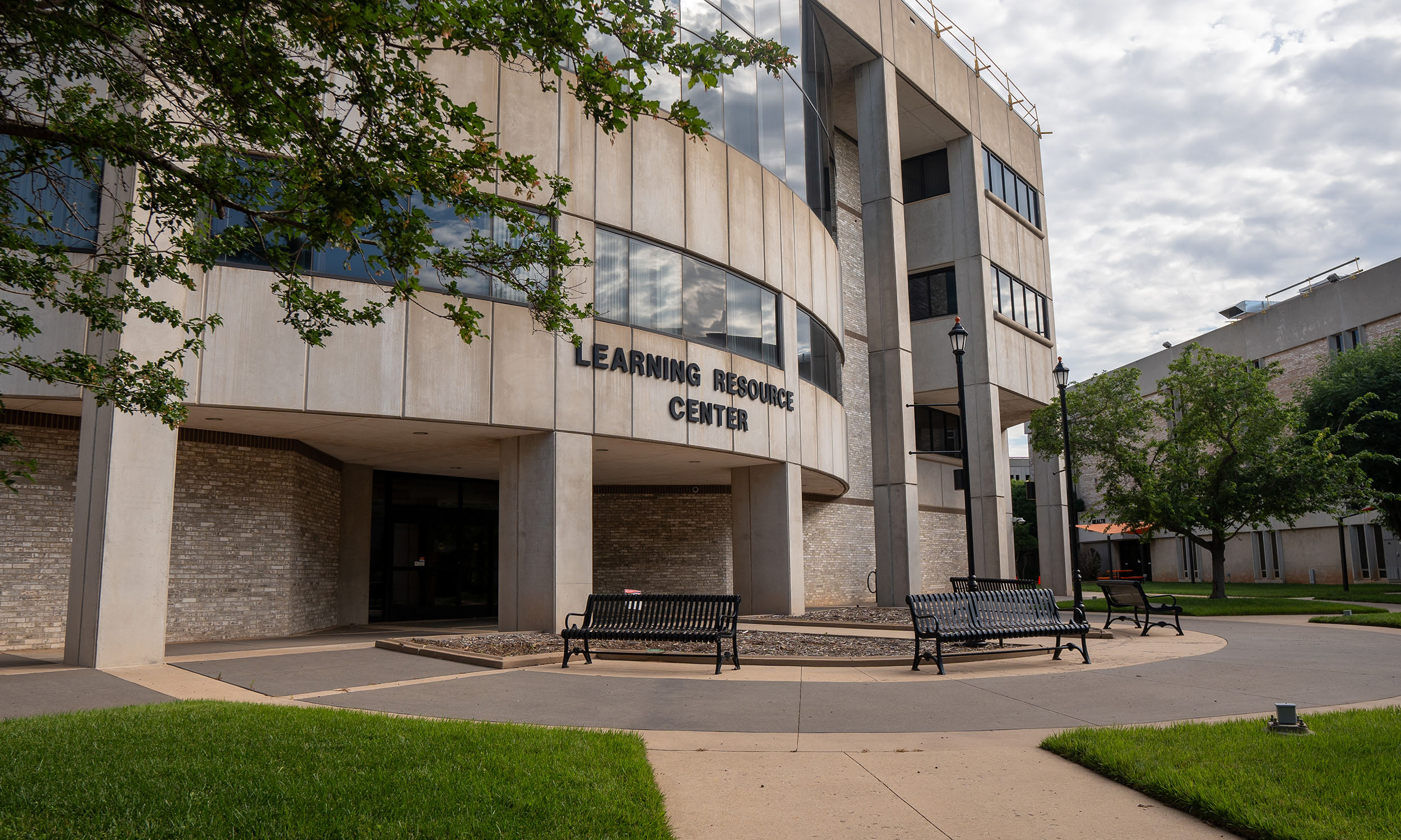 The exterior of a modern building labeled 'Learning Resource Center' featuring large windows and a curved facade. Benches and a lamp post sit near the entrance, surrounded by neatly maintained grass and trees.