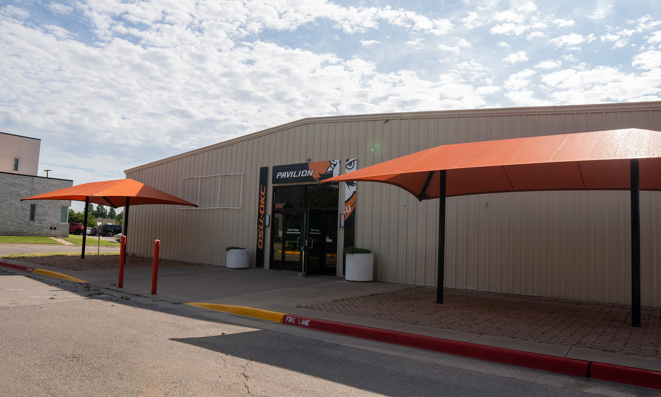 A building with a beige exterior and a sign above the entrance that reads 'Pavilion.' Two large orange canopies on either side of the entrance provide shade over the sidewalk. The entrance has glass doors with a small sign on them.