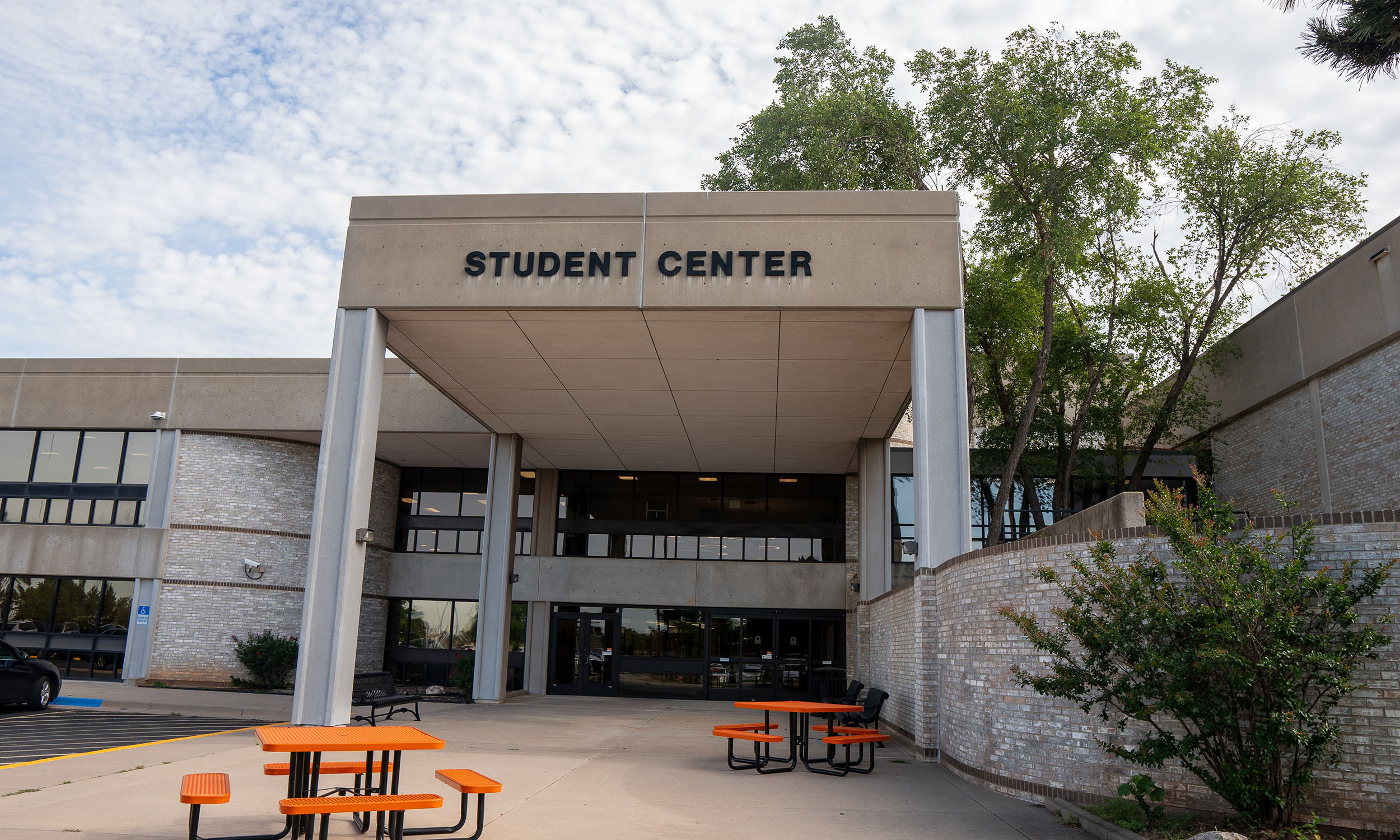 Entrance to a modern building labeled 'Student Center' with large windows and concrete pillars. Several orange picnic tables and benches are arranged on a paved area in front, surrounded by trees and greenery under a partly cloudy sky.