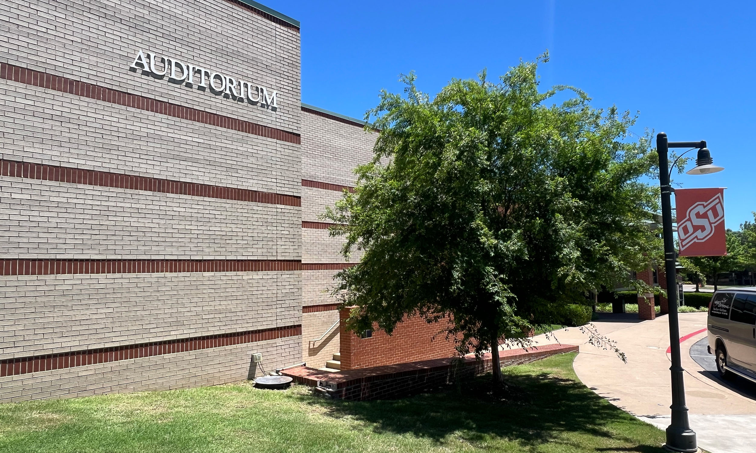 The exterior of a building with the word 'AUDITORIUM' displayed on it. A tree stands in front of the building, and a lamp post with an Oklahoma State University banner is visible. The sky is clear and blue, with grass in the foreground.