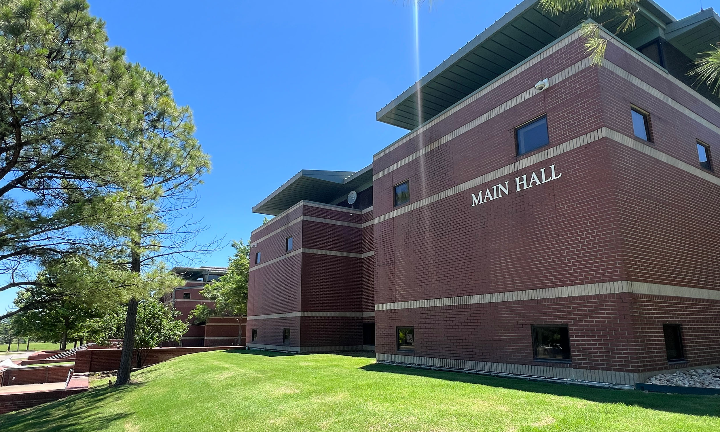A large brick building labeled 'Main Hall' with a green lawn and trees in the foreground. The sky is clear and blue.