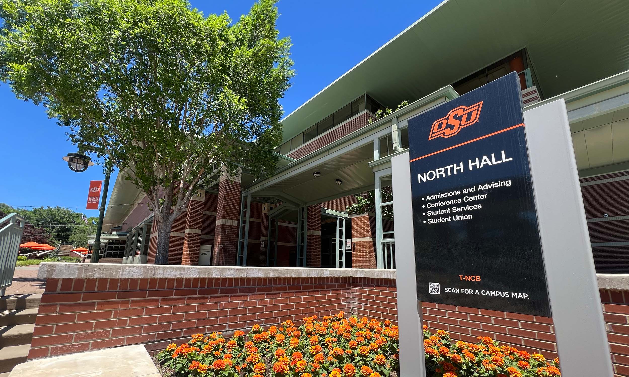 The exterior of North Hall at Oklahoma State University. A sign in the foreground lists services available in the building, including Admissions and Advising, Conference Center, Student Services, and Student Union. The building is surrounded by a well-maintained garden with orange flowers and trees. Outdoor seating areas with orange umbrellas are visible in the background.