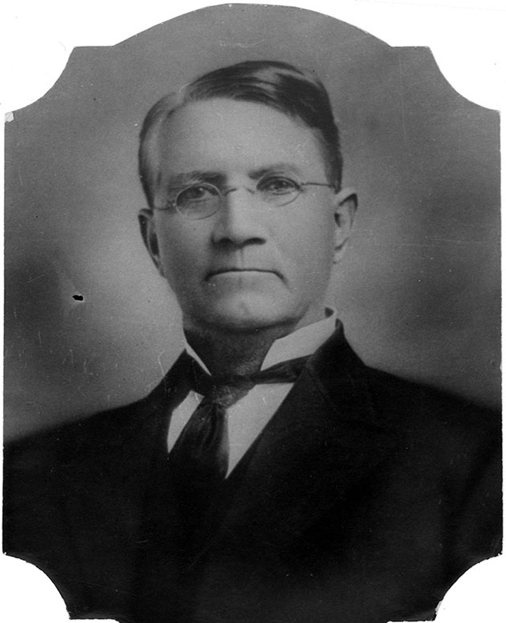 A black and white portrait of a man wearing a suit and tie. The background is plain and dark, with the edges of the image having an ornate cut-out design.