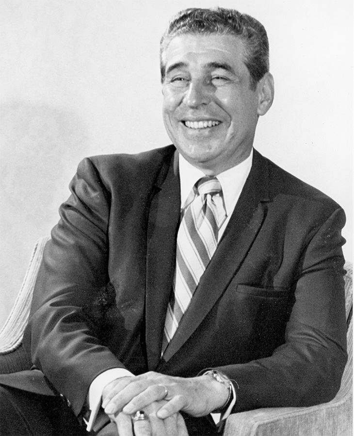 A man wearing a dark suit, white dress shirt, and striped tie, seated on a chair with hands clasped in front. 