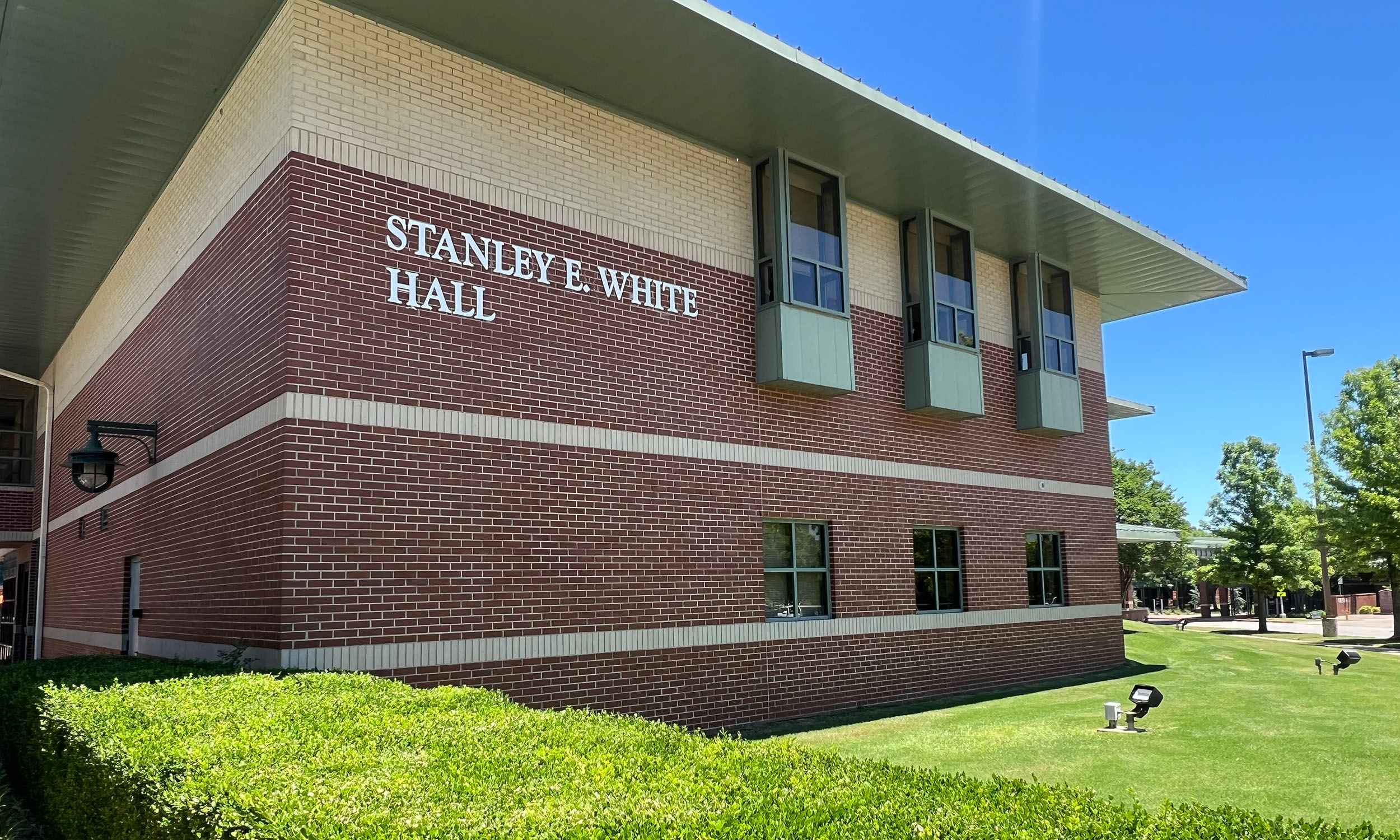 A two-story brick building with the name 'Stanley E. White Hall' displayed on the upper left side of the facade. The building has large windows and a sloped roof. There is a well-maintained lawn with bushes in front of the building, and trees are visible in the background under a clear blue sky.