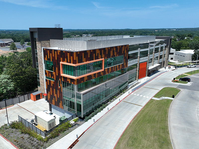 A modern multi-story building with a facade of orange, brown and black panels. It features large glass windows and an adjacent parking area with a curved driveway. Trees and other buildings are visible in the background under a clear blue sky.