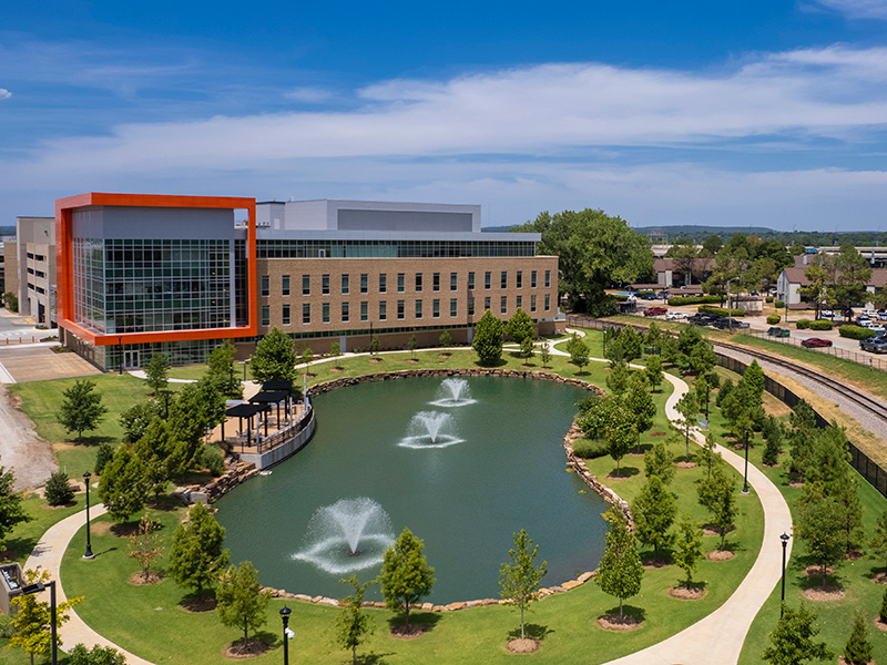 Aerial view of a modern building with a large glass facade and an orange frame on the left. In front is a landscaped area with a pond containing three fountains, surrounded by walking paths and trees. Additional buildings and greenery are visible under a clear blue sky.