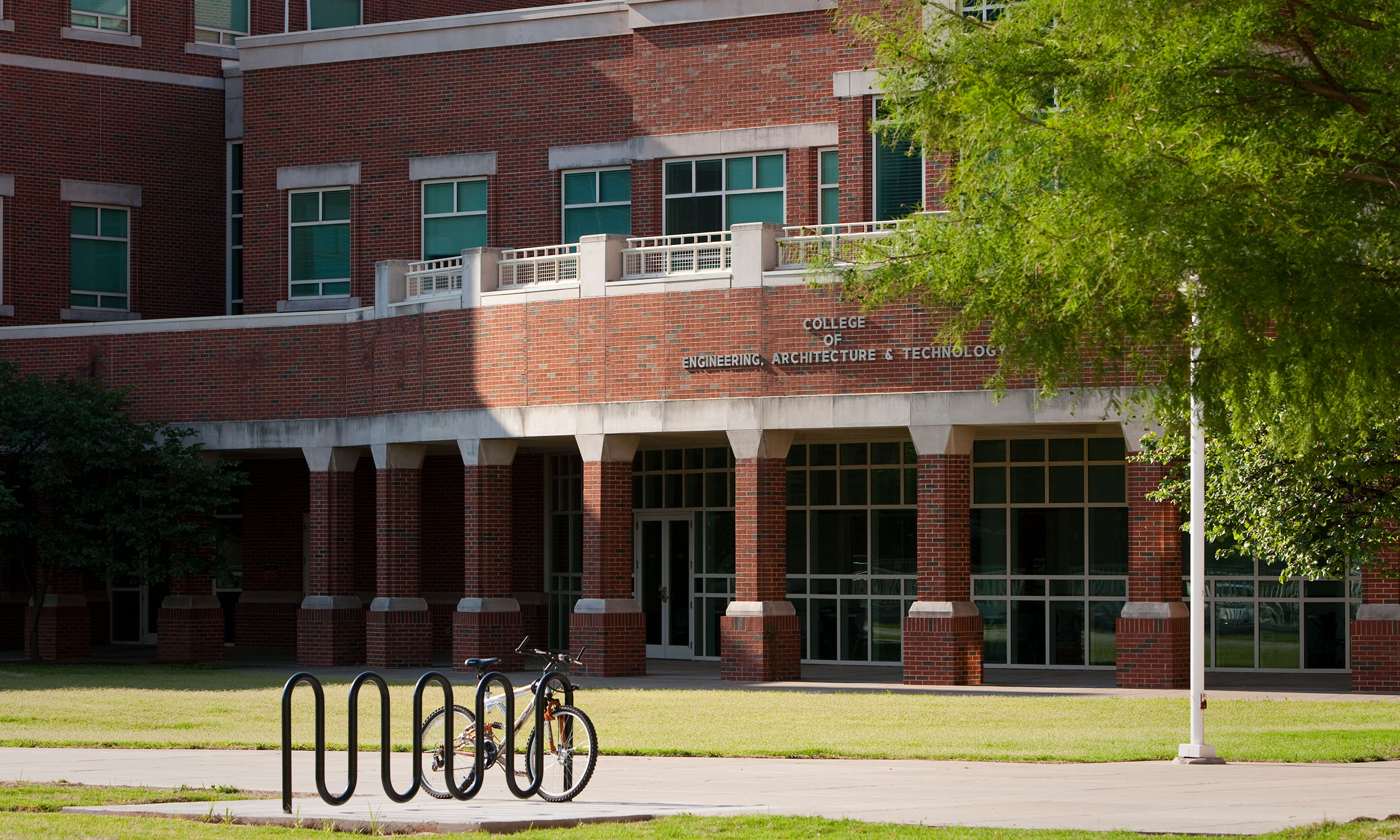 A brick building with large windows and a sign that reads 'College of Engineering, Architecture & Technology' above the entrance. There is a bicycle parked at a bike rack in front of the building, and a tree with green leaves partially shades the area. 