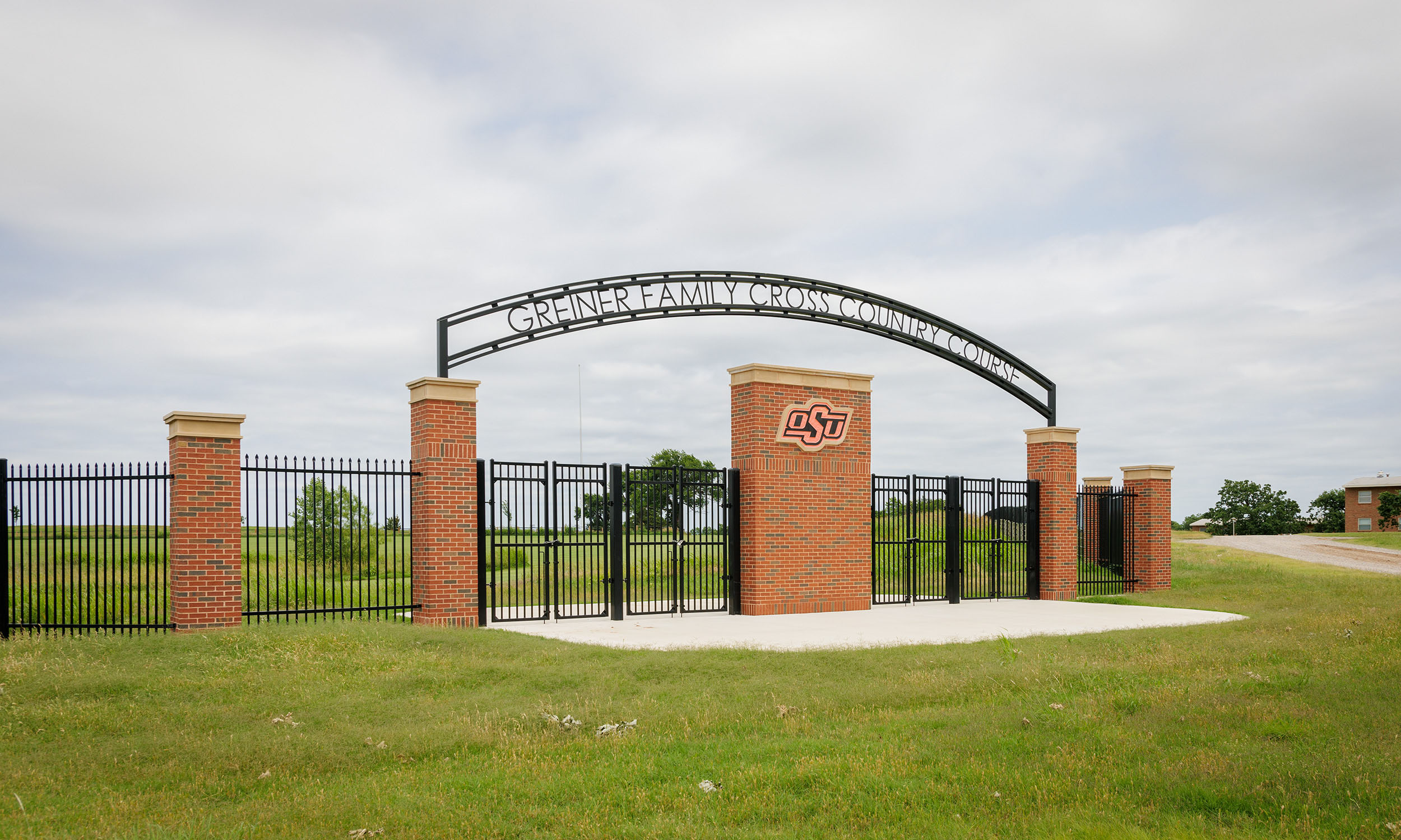 The entrance gate to the Greiner Family Cross Country Course at Oklahoma State University. The gate is made of black metal fencing with brick pillars and an archway that has the name of the course written on it. The OSU logo is displayed on one of the central brick pillars. 